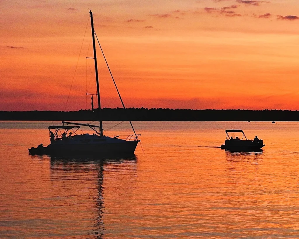 Sailboat and pontoon boat silhouetted on a lake during a vibrant orange sunset.